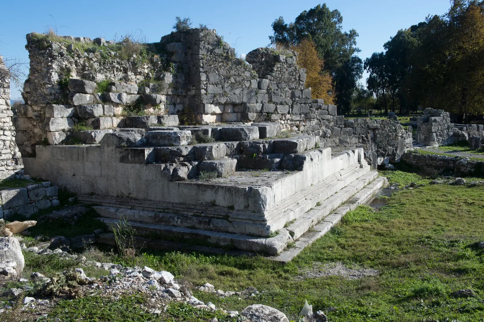 Limyra antik kentinde ağaçlar arasında taş kalıntılar ve arkasında yükselen kayalık yamaç